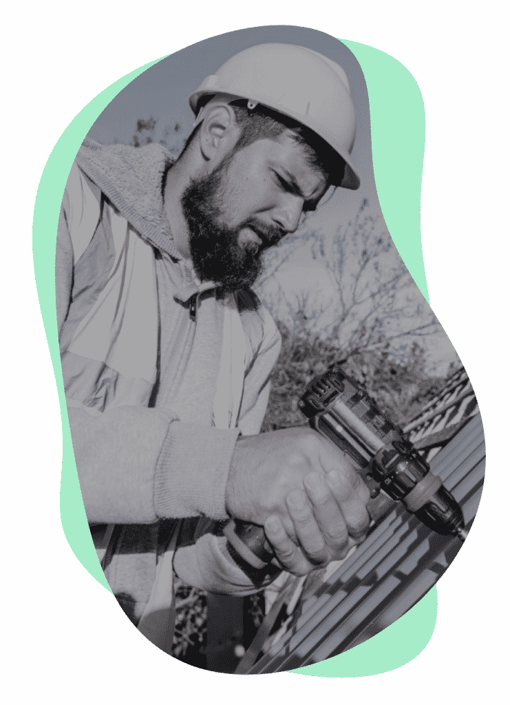 A construction worker wearing a hard hat and safety vest is using a power drill while installing roofing materials. The background features trees and a partially completed structure. The image is predominantly in black and white, with a green abstract shape framing it.