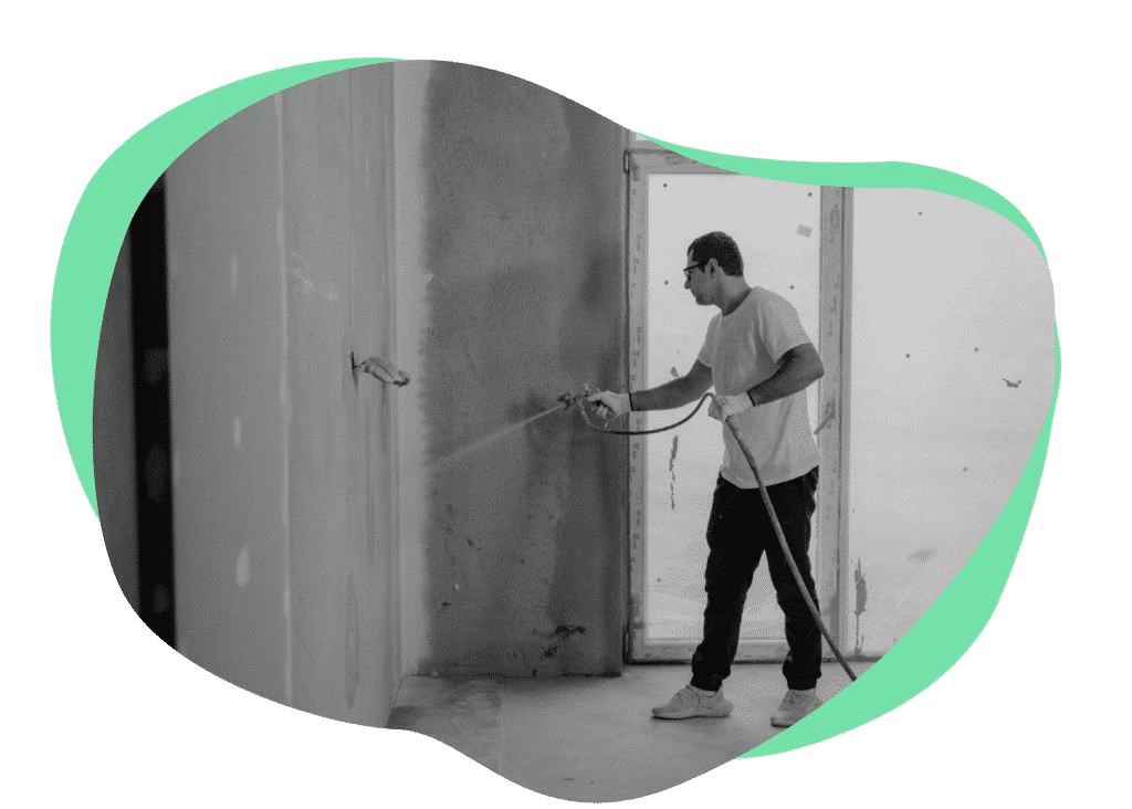 A man is using a spray gun to paint a wall in an unfinished room. He is wearing a white t-shirt and black pants, and protective eyewear. The background shows a window and bare walls with some construction debris.